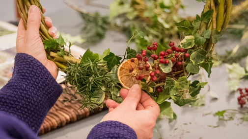Close-up of hands in a blue sweater creating a decorative wreath. The wreath features fresh green foliage, red berries, rosemary sprigs, and a dried orange slice being attached to the arrangement."